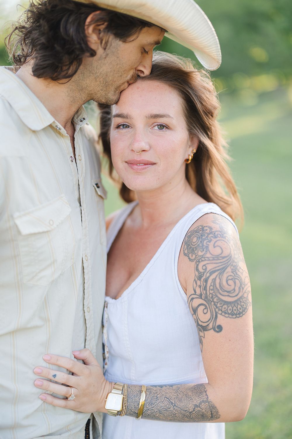 A romantic outdoor portrait shows a couple in casual attire, with one wearing a white hat and the other displaying ornate arm tattoos.