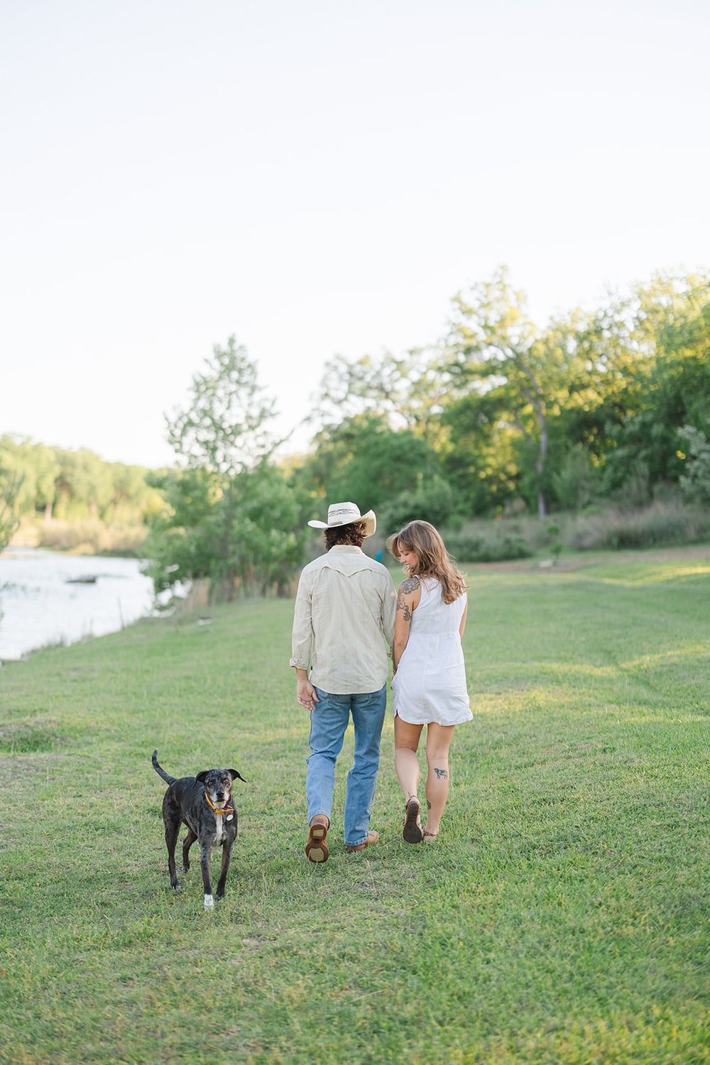 A couple walking their dog along a grassy riverside path during a golden sunset.