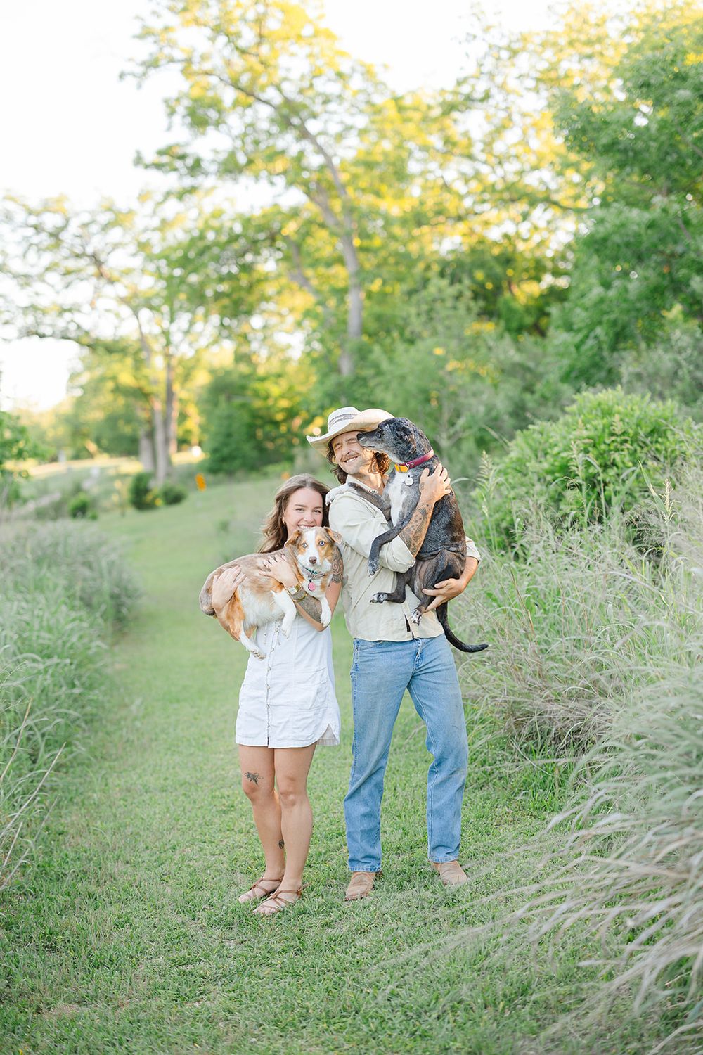A couple enjoys a sunny afternoon outdoors with their small dogs on grassy terrain with trees in the background.