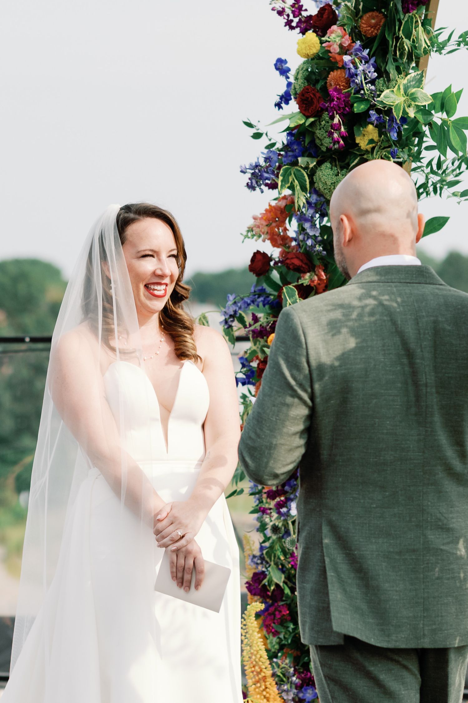 A joyful wedding ceremony takes place under a vibrant floral arch with colorful blooms of purple, red and yellow flowers.
