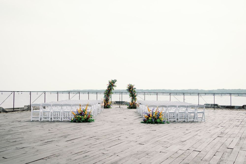Empty white chairs lined up on a wooden boardwalk facing a calm beachfront under an overcast sky.