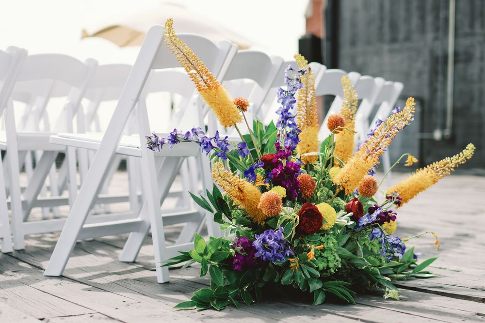 Low floral centerpiece with orange birch branches and purple flowers surrounded by white garden chairs at an outdoor wedding.