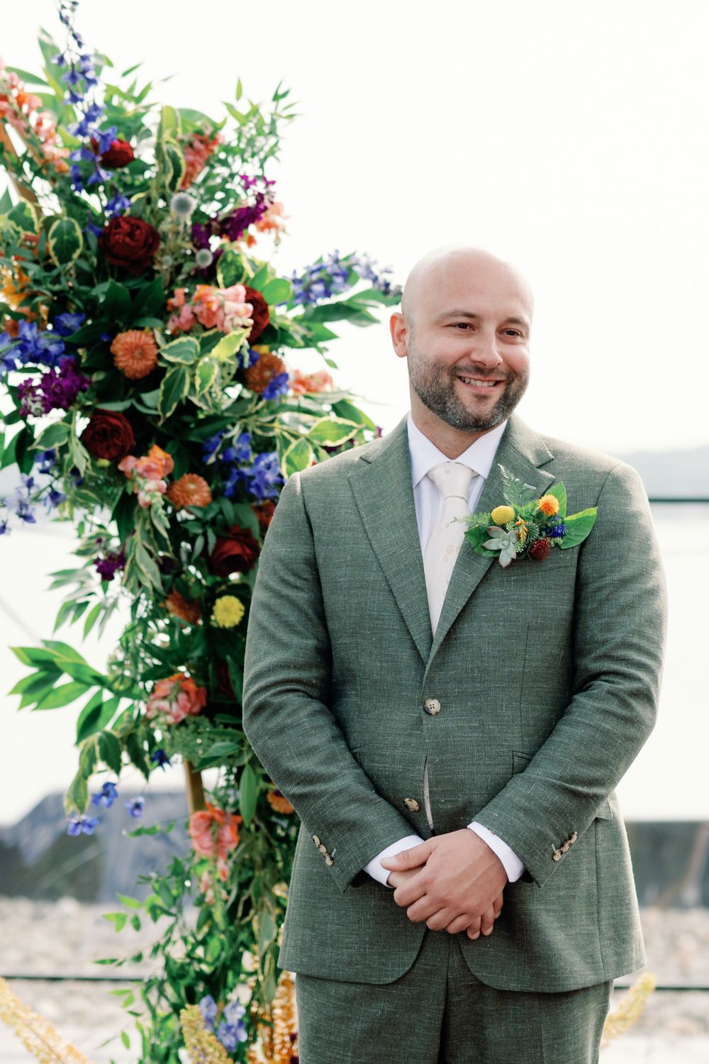 Wedding ceremony decorated with vibrant floral arch featuring red, peach, and blue blooms alongside a green suit.