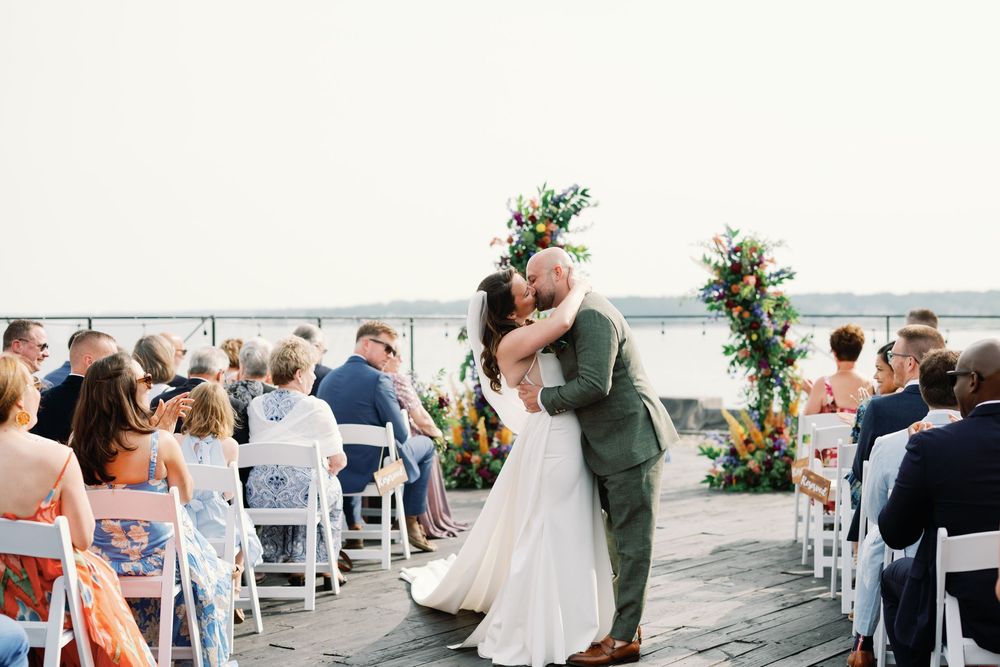 A romantic wedding ceremony on a waterfront deck with a floral arch and white chairs overlooking the water on a sunny day.