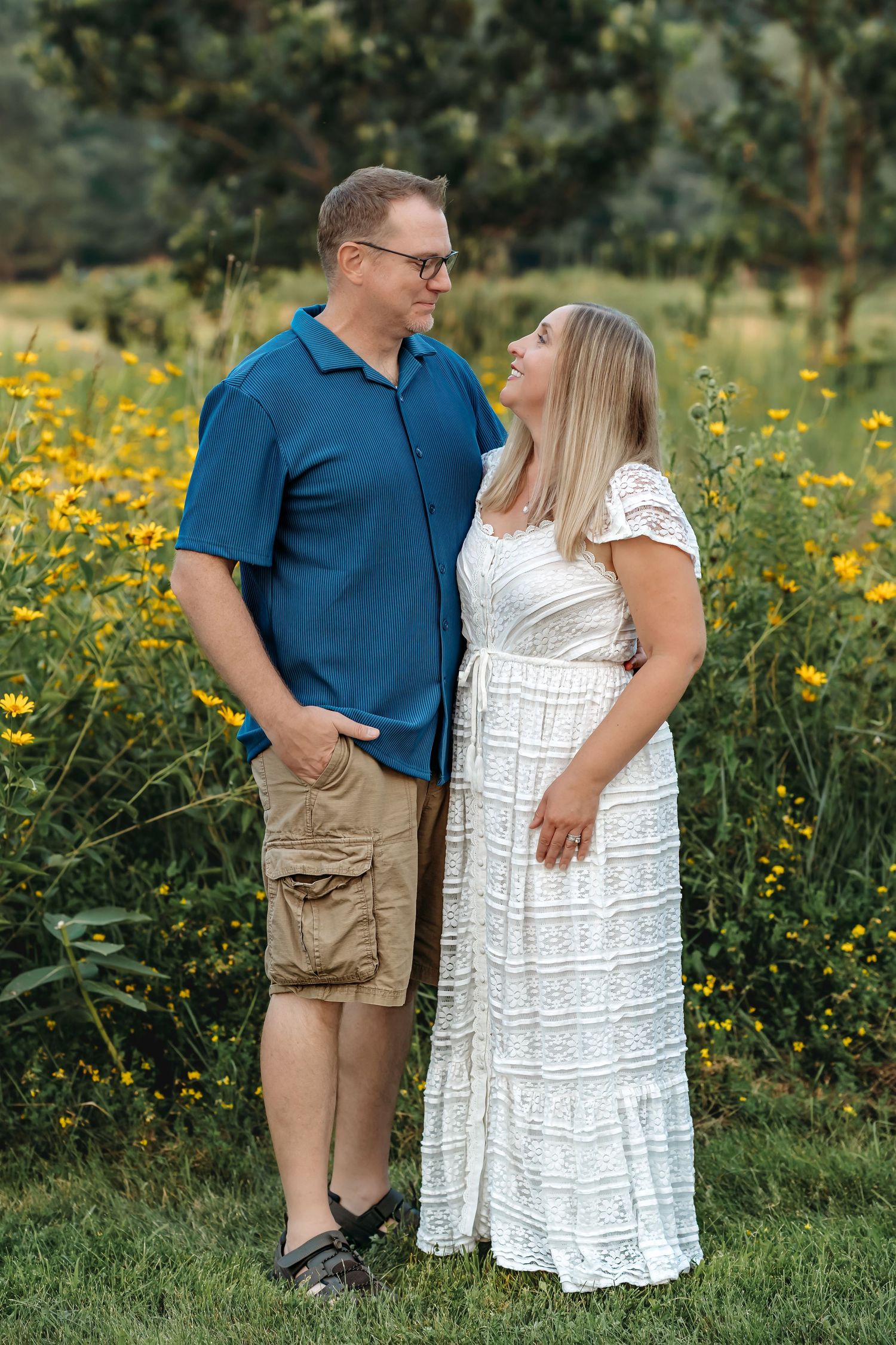 A couple poses lovingly in a field of yellow wildflowers, with her wearing a white dress and him in a blue shirt.