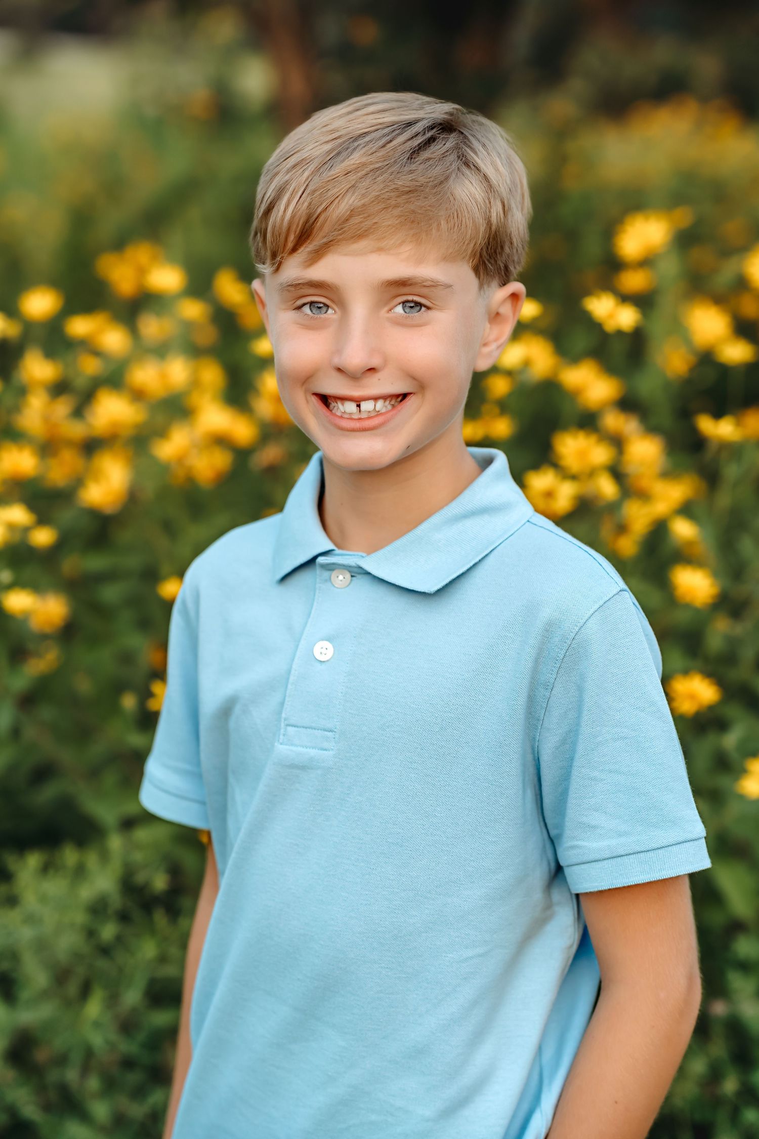 Child wearing a light blue polo shirt smiles brightly in front of yellow flowers on a sunny day.
