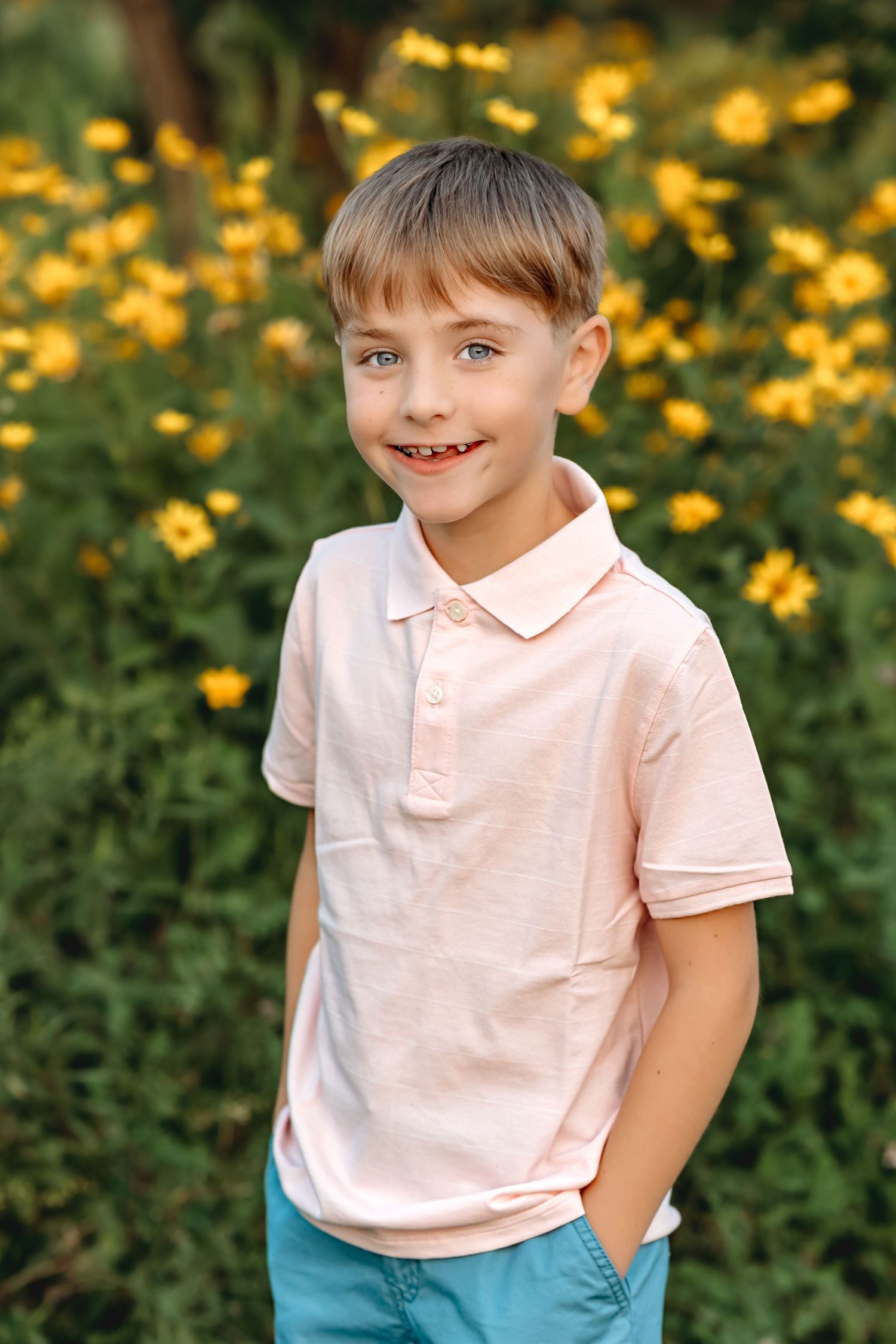 Child wearing pink polo shirt and teal shorts poses against yellow wildflowers in garden setting.