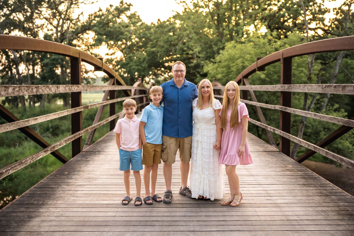 A family of four stands together on a wooden bridge during a summer evening photoshoot.