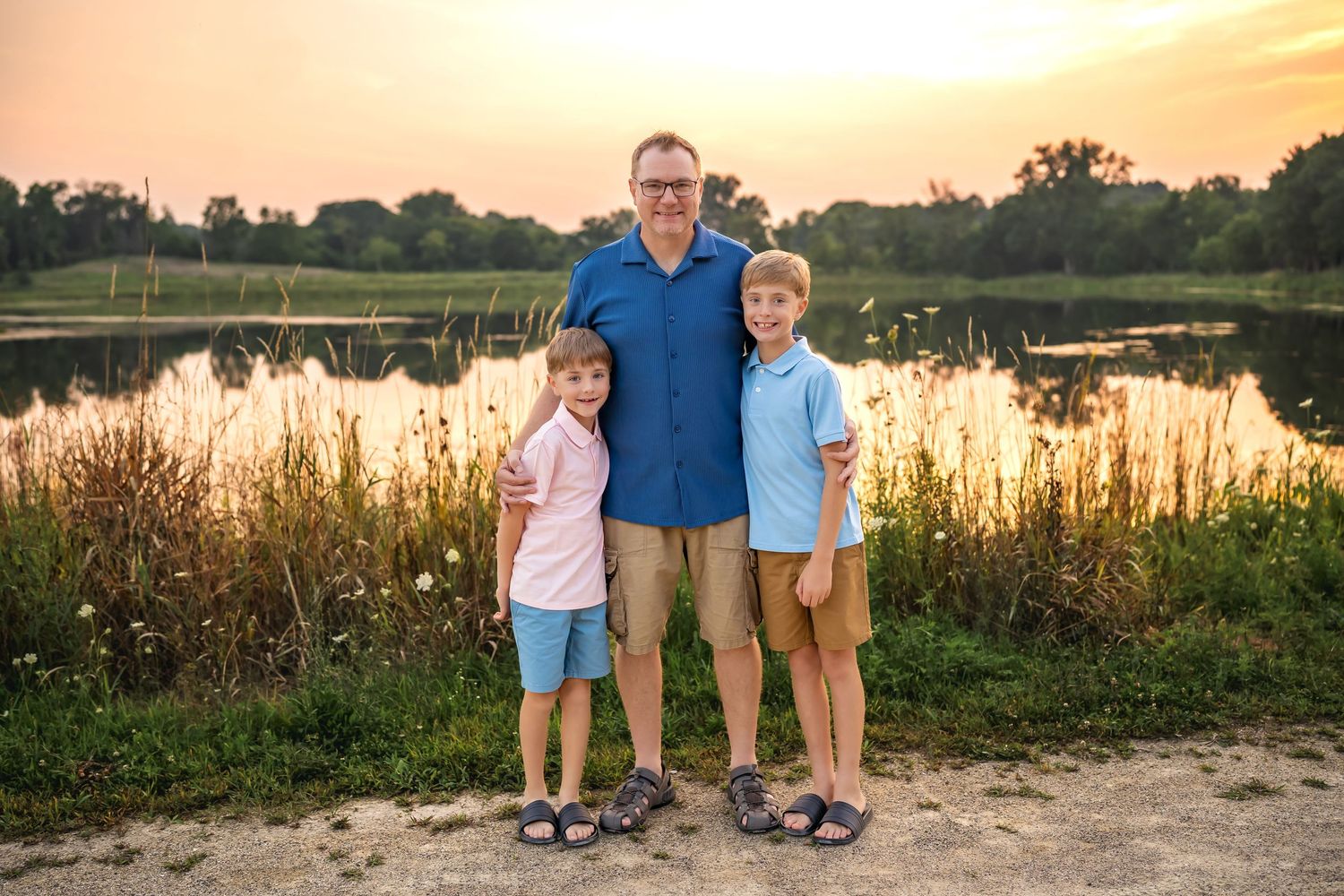 Family standing by lake at golden hour with tall grass and glowing sunset in background.