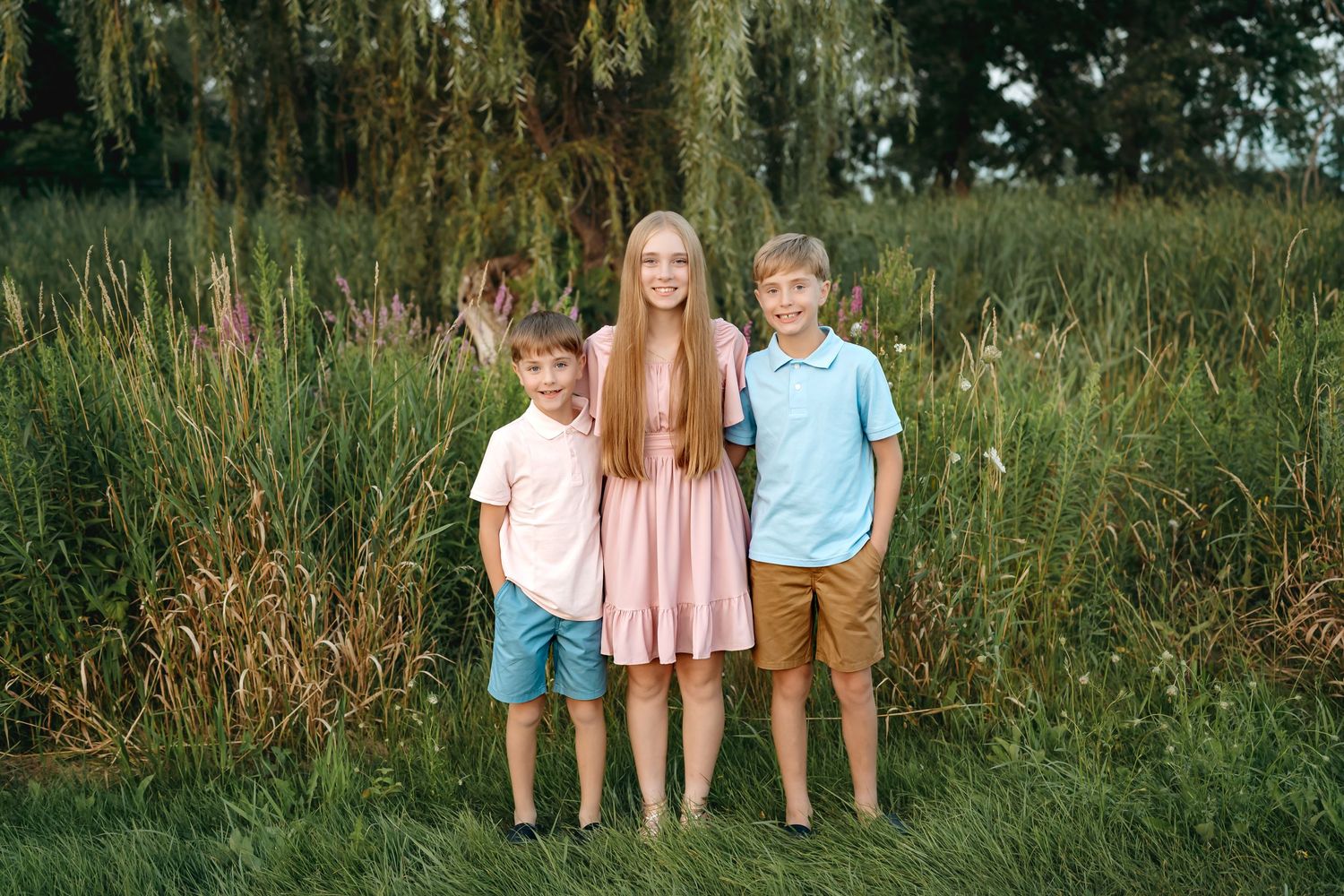 Three children stand together amidst tall grass and yellow wildflowers in a natural outdoor setting.