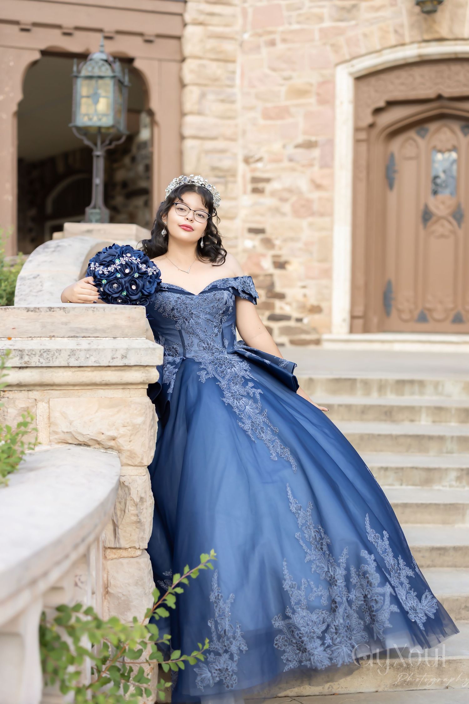 A person in an elegant navy blue off-shoulder ball gown with lace details poses on stone steps near a vintage doorway.