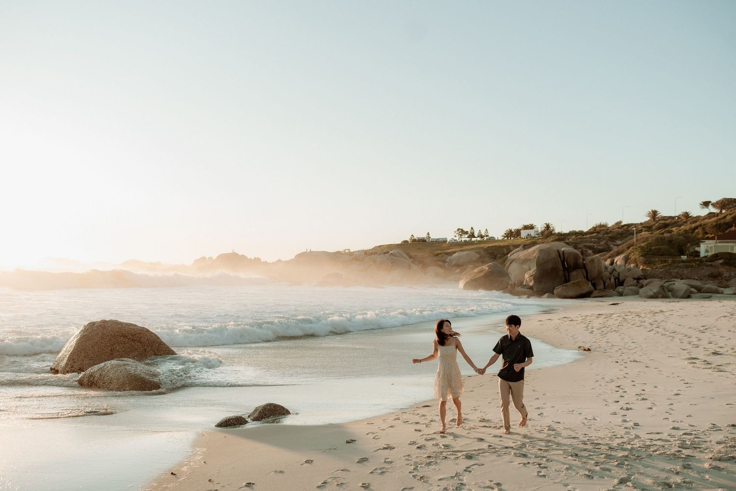 Couples walk along a misty sandy beach at sunset with waves crashing against rocky outcrops in the background.