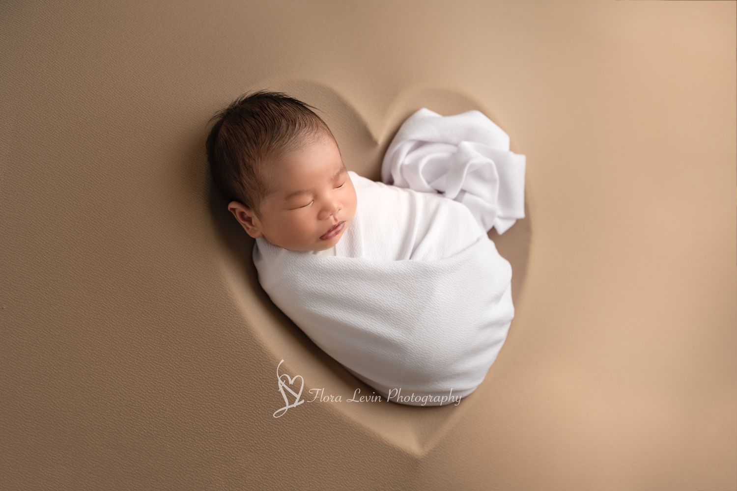 A newborn baby swaddled in white fabric lies peacefully against a neutral brown background in the shape of a in a professional studio photo.