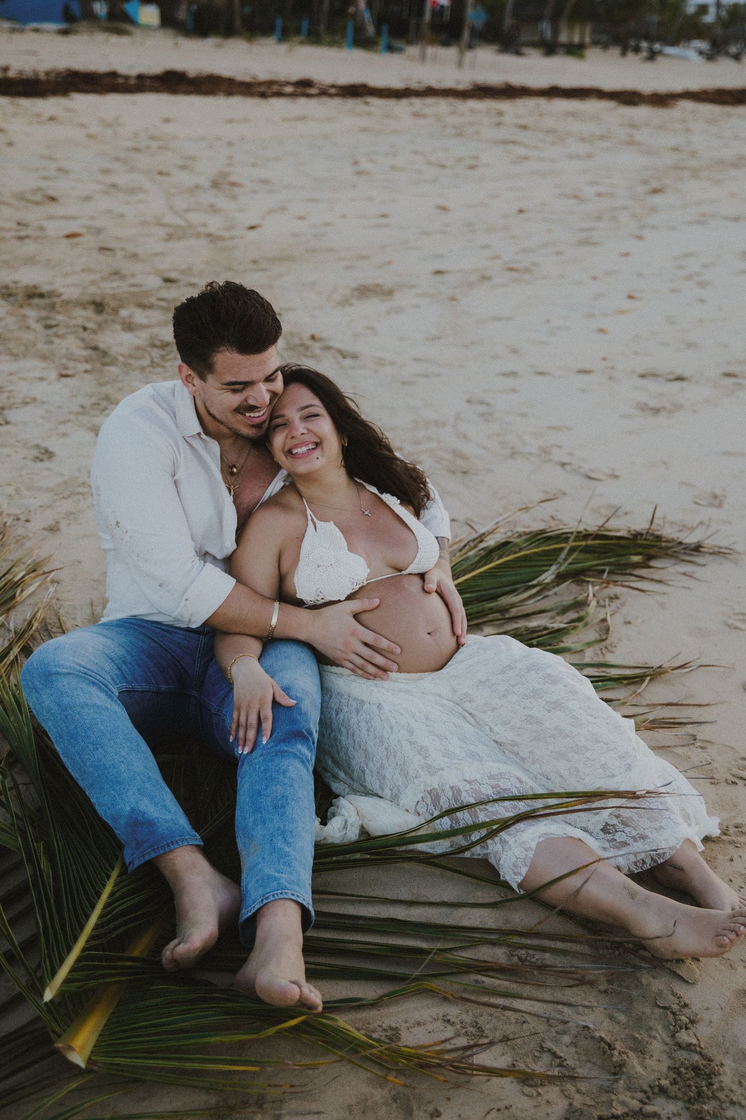 Romantic beach photoshoot with couple lounging on palm fronds by the ocean.