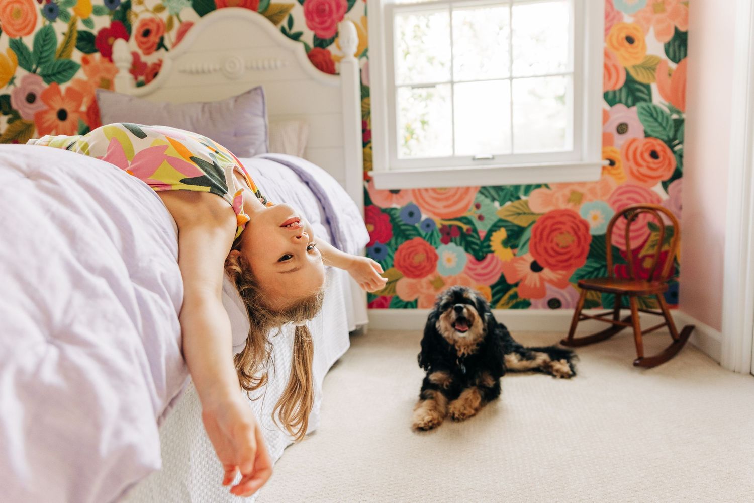 A joyful moment in a colorful floral wallpapered bedroom with a dog resting near a rocking chair.