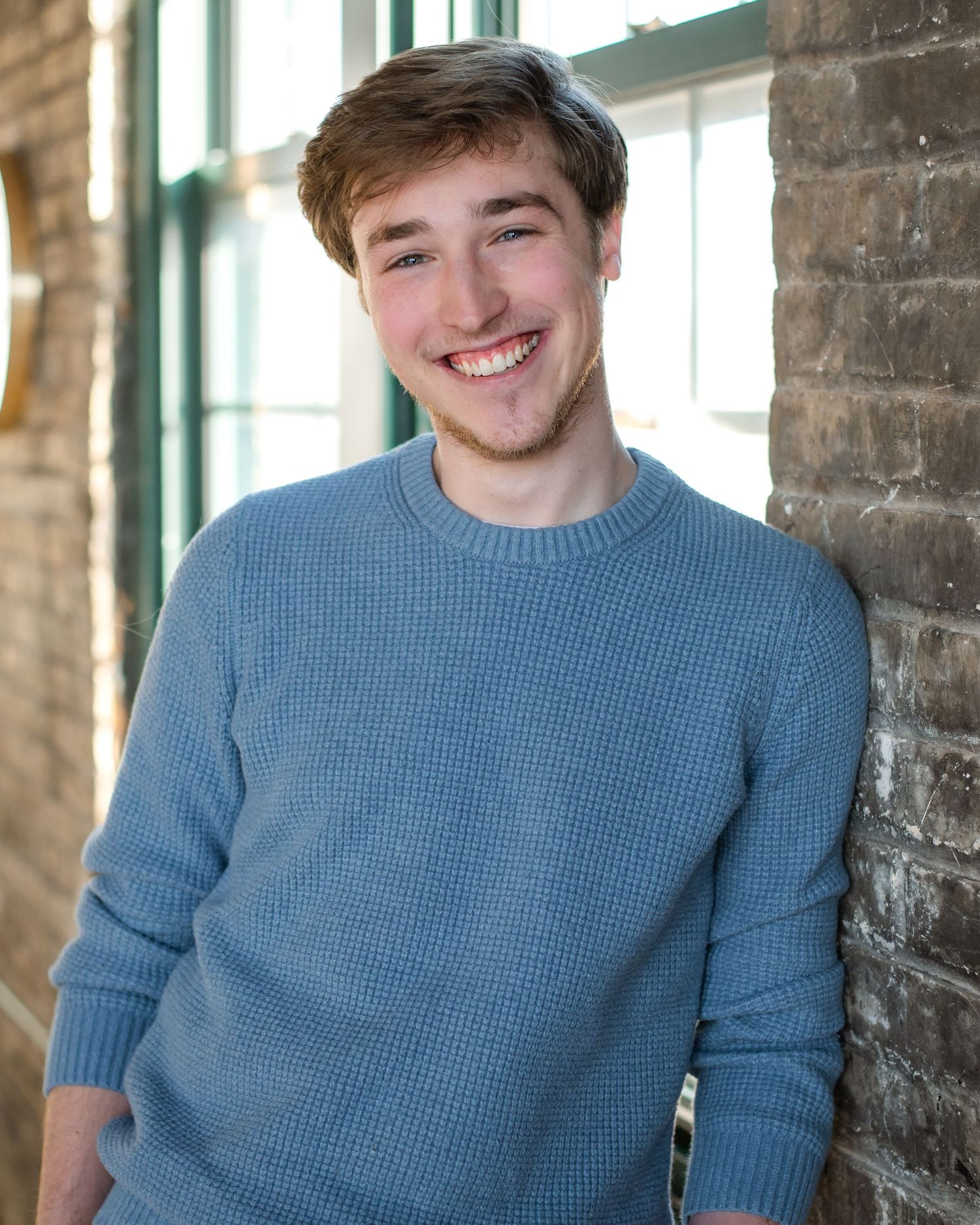 A person in a light blue knit sweater smiles warmly while standing against a brick wall near a window.