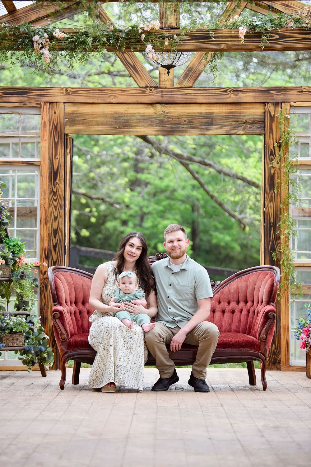 A young family with a baby poses on a red tufted sofa in a rustic wooden greenhouse surrounded by lush greenery.