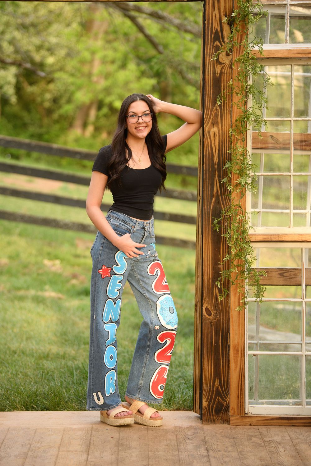 A smiling teenage girl poses in painted senior 2026 jeans and black top beside a rustic wooden door with greenery.