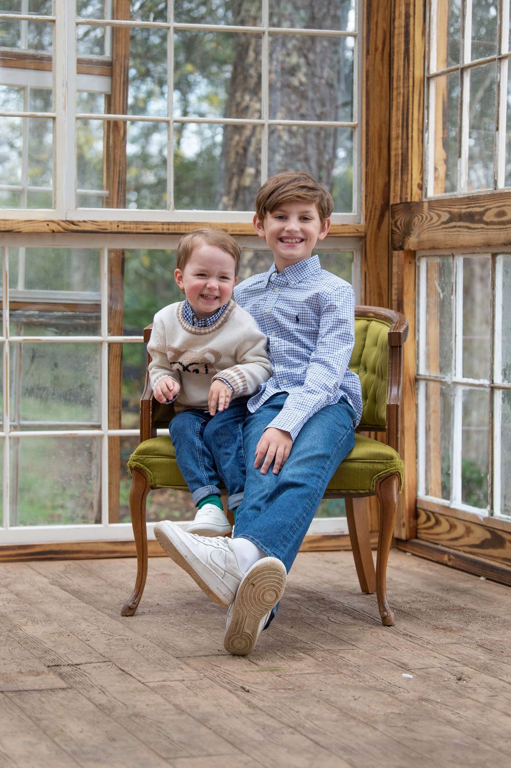 Two children laugh together while sitting in wooden chairs on a rustic porch with large windows.