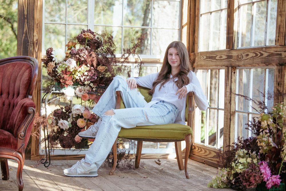 Wedding couple lounges near elegant floral arrangements in a light-filled rustic setting with tall windows.
