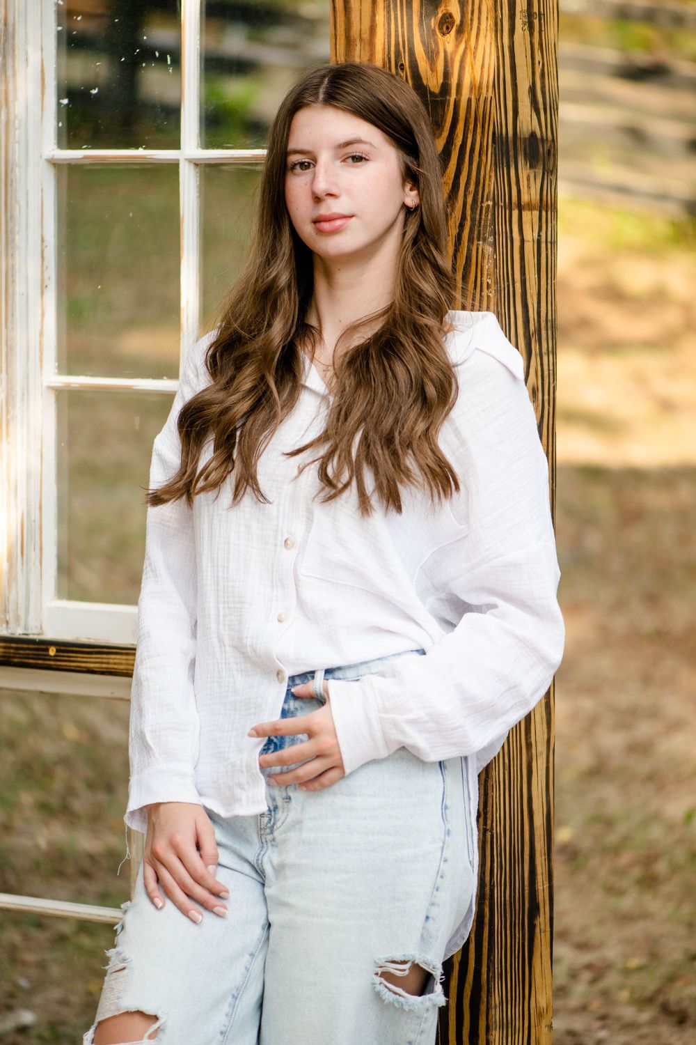 Casual outdoor portrait of someone wearing a white button-down shirt and light wash jeans leaning against a wooden post.