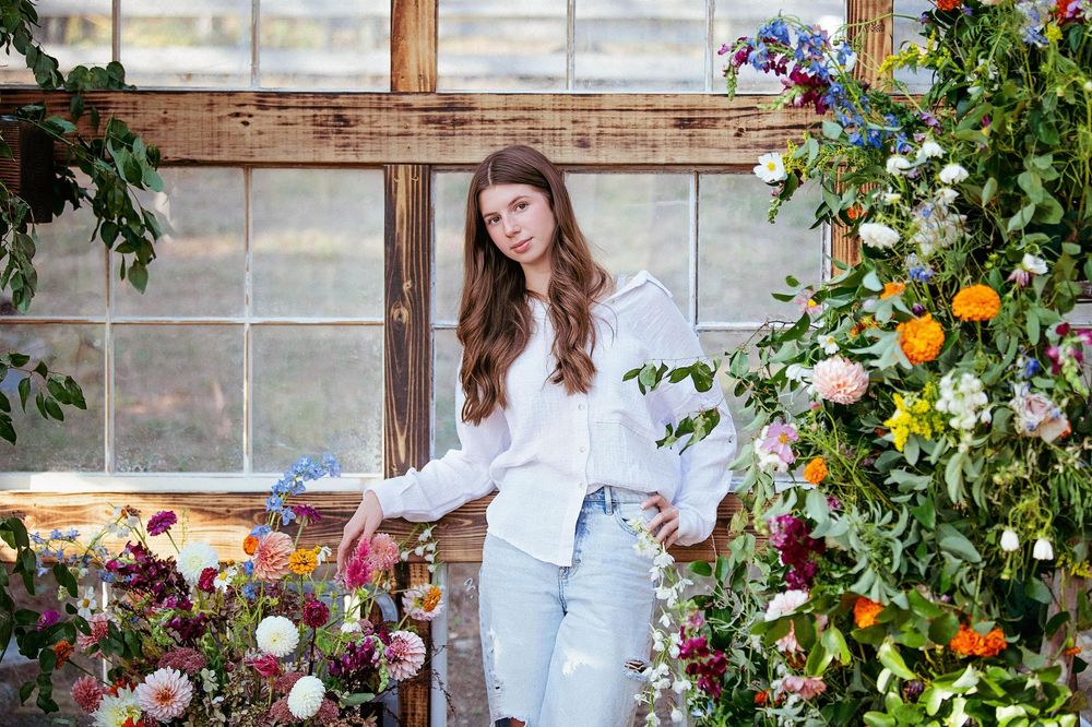 A greenhouse worker tends to blooming flowers and plants in a rustic wooden greenhouse filled with natural light.