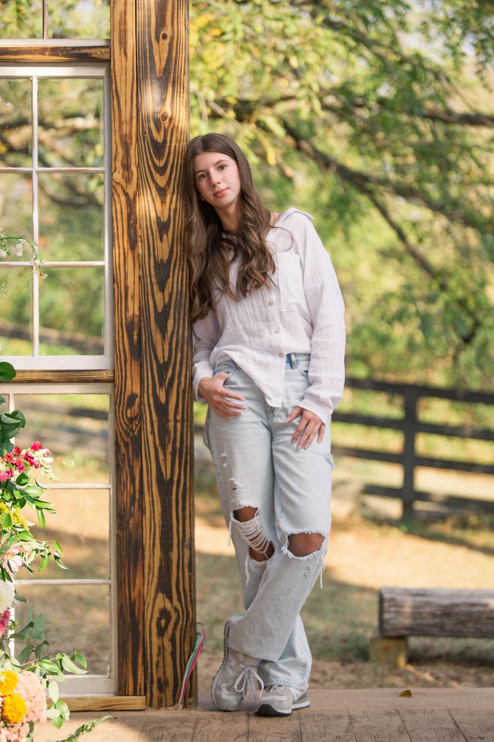 Casual outdoor portrait with white sweater and distressed jeans against rustic wooden post and garden setting.
