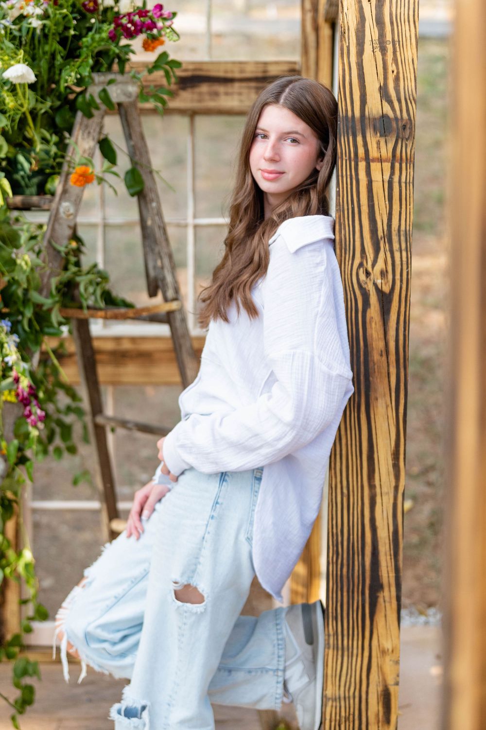 A person in a white oversized shirt and distressed light blue jeans poses against a wooden pillar with flowering vines.