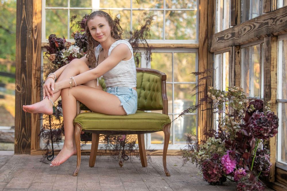 A rustic sunroom with wooden beams features someone relaxing on a green cushioned chair beside potted purple flowers.