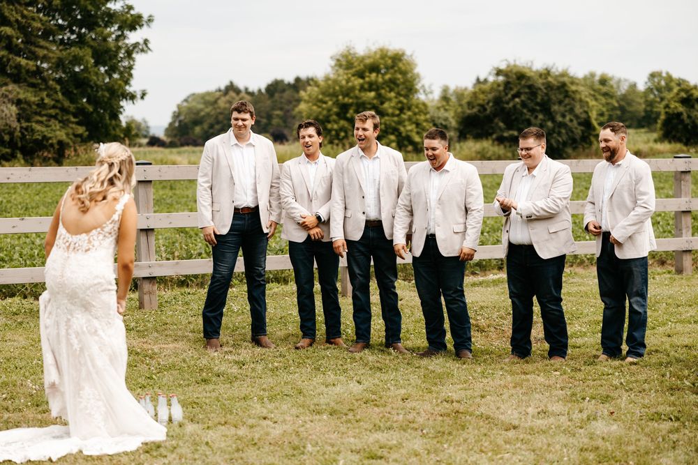 Wedding party members celebrate and pose together along a white fence in a sunny outdoor setting.