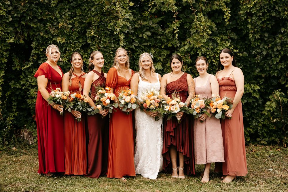 Wedding party wearing burgundy and white dresses poses together against lush green garden backdrop.