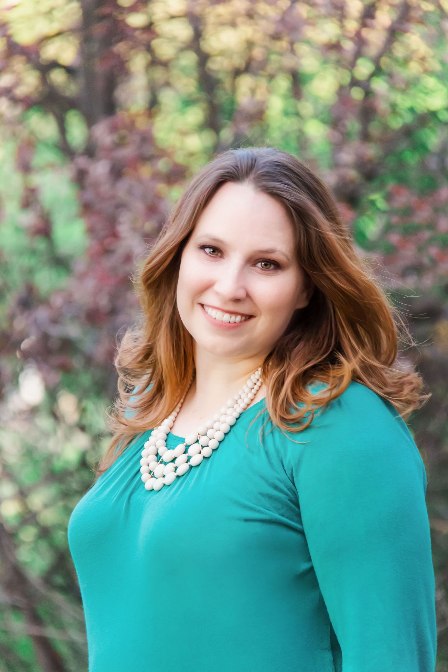 Woman in teal blouse with pearl necklace posing against blurred natural background.