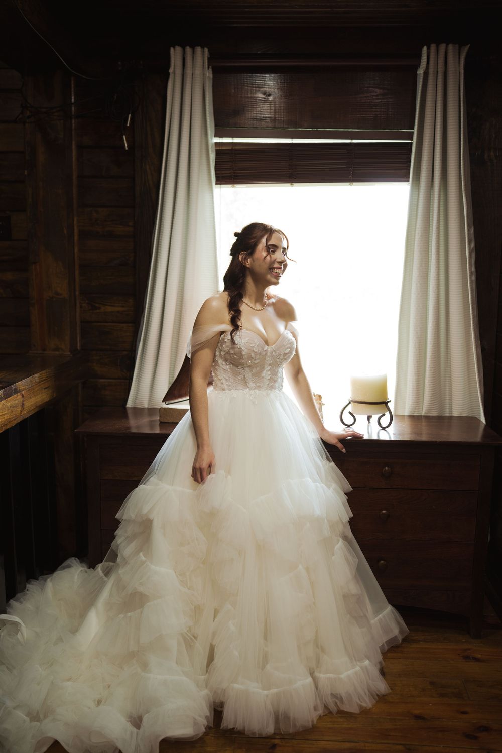 Bride in voluminous tulle wedding dress poses by rustic cabin windows with natural lighting.