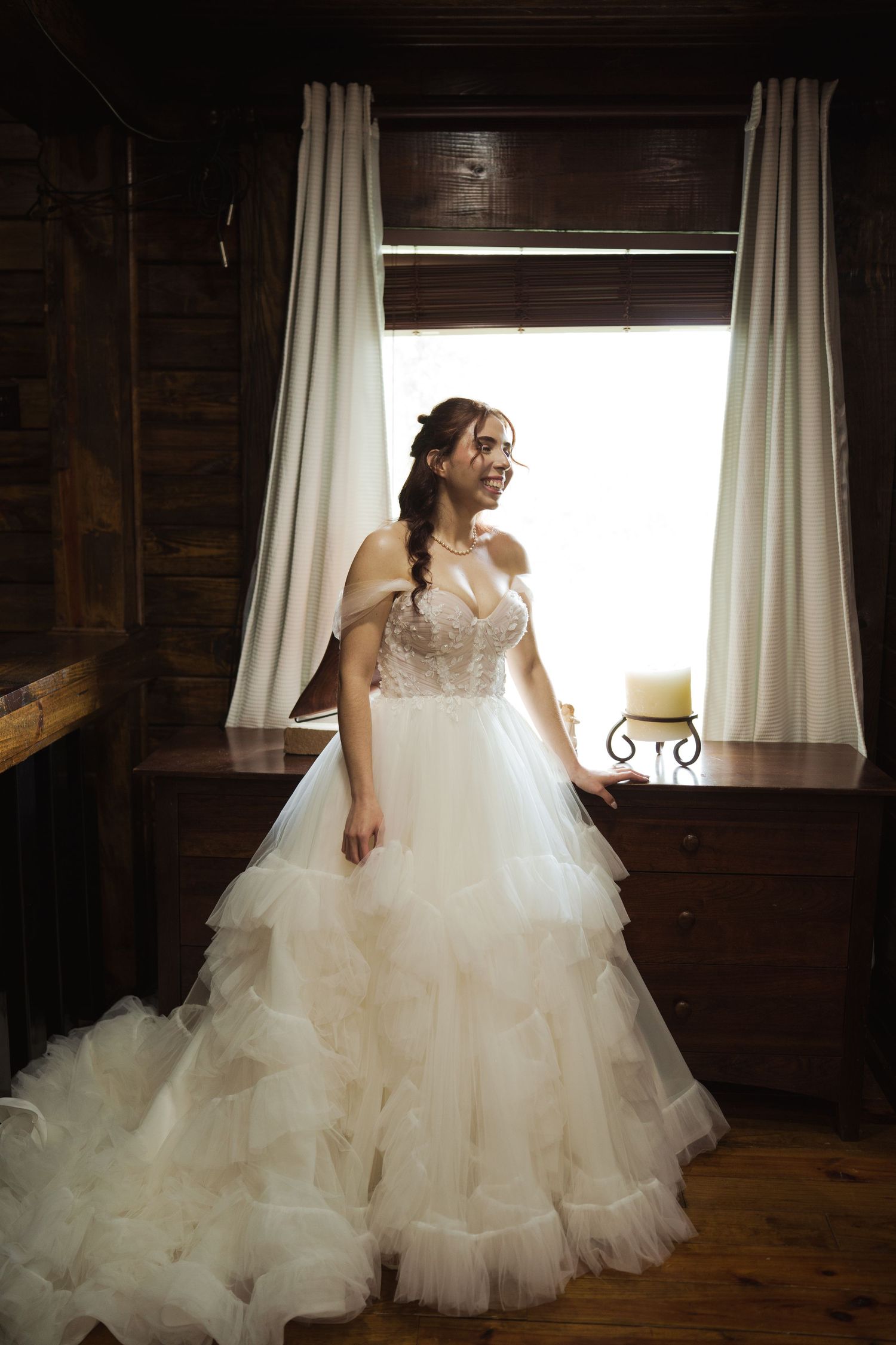 Bride in voluminous tulle wedding dress poses by rustic cabin windows with natural lighting.