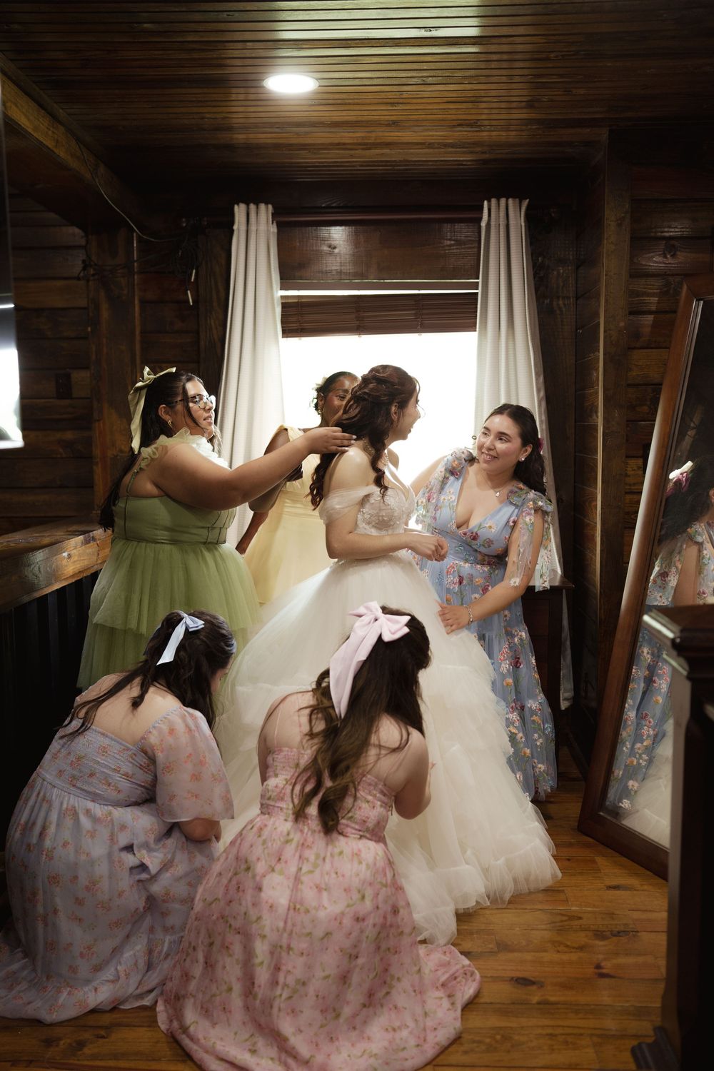 Bridesmaids in floral dresses help adjust the bride's gown in a rustic wooden room.