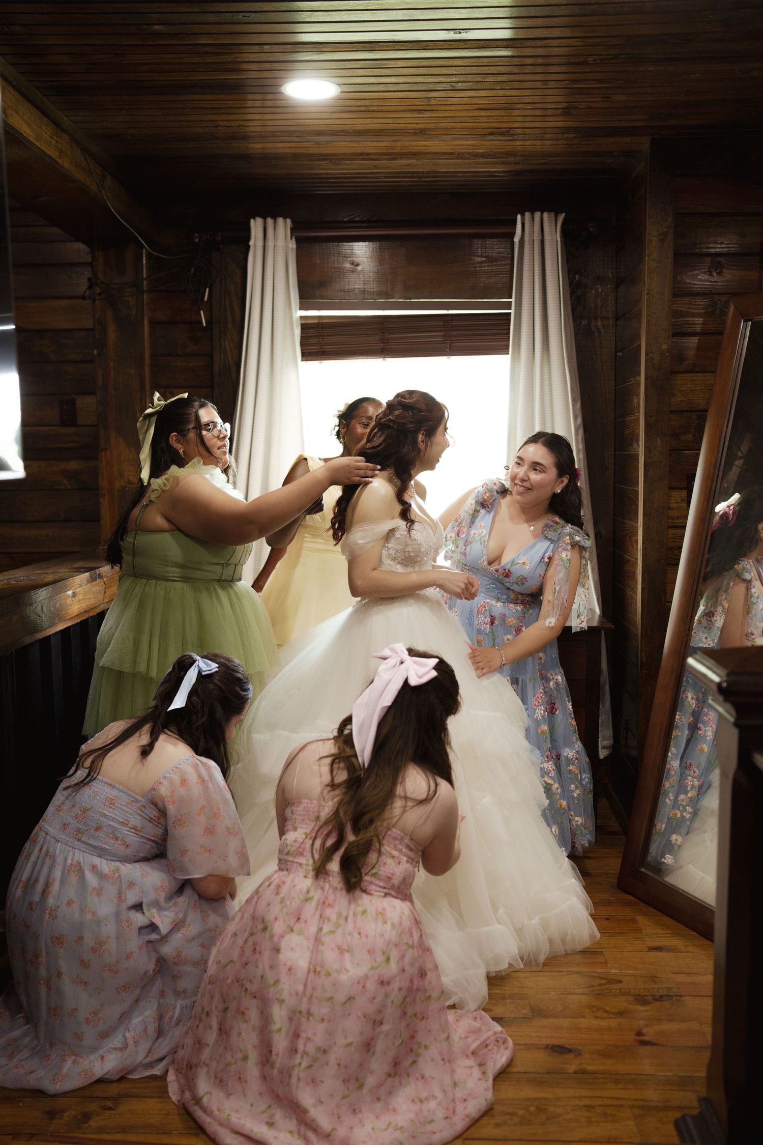 Bridesmaids in floral dresses help adjust the bride's gown in a rustic wooden room.