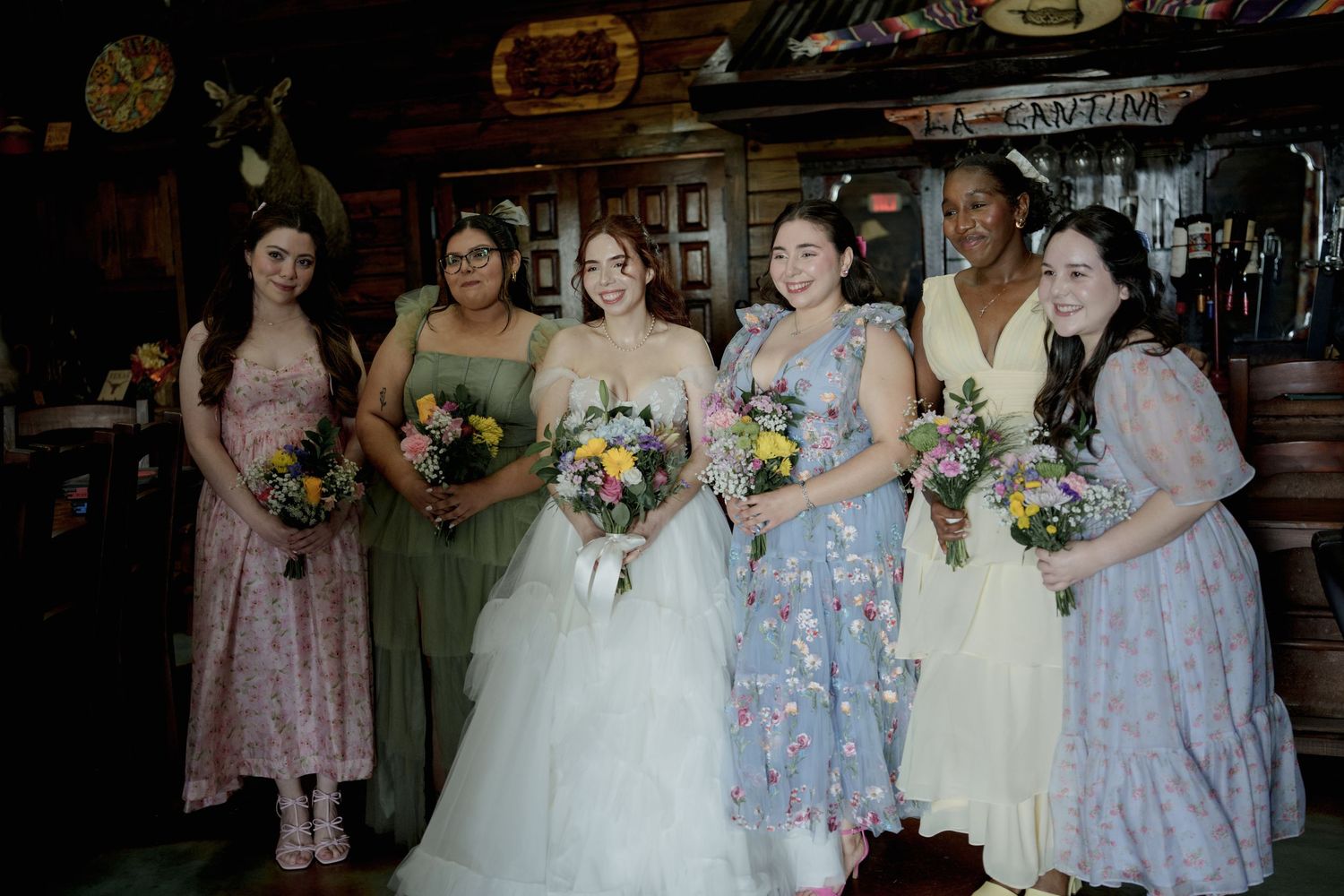 Bridesmaids in pastel floral dresses gather for photos at rustic wedding venue.