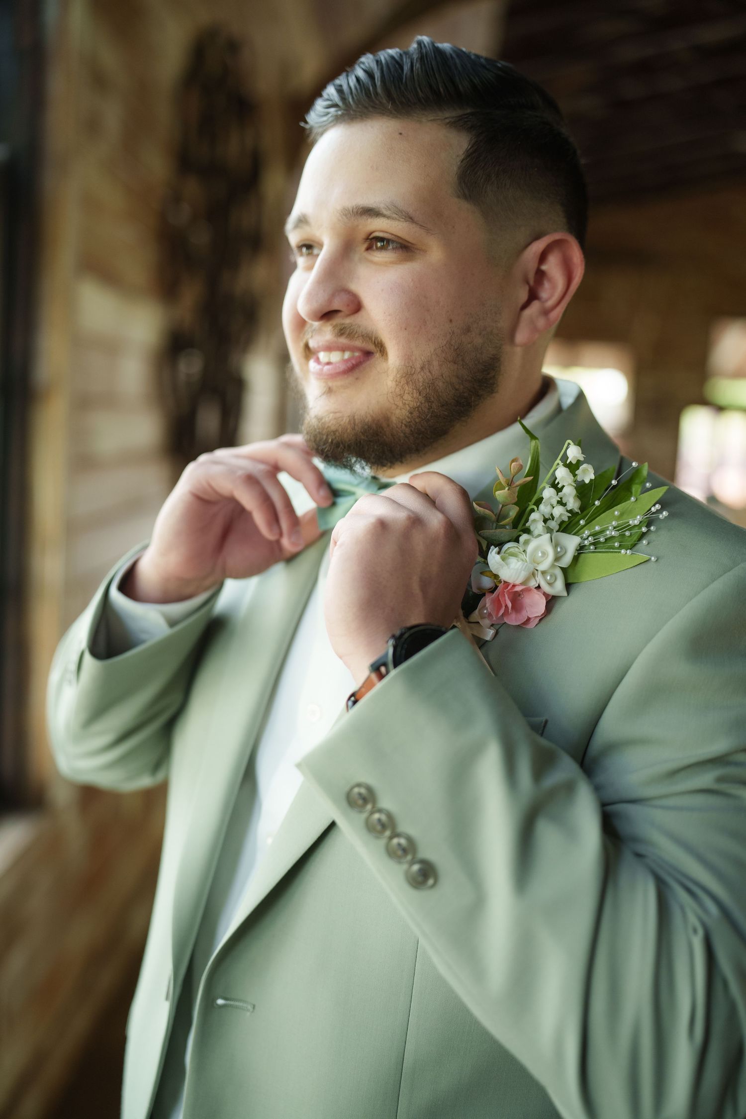 Groom in mint green suit jacket adjusts boutonniere while smiling.