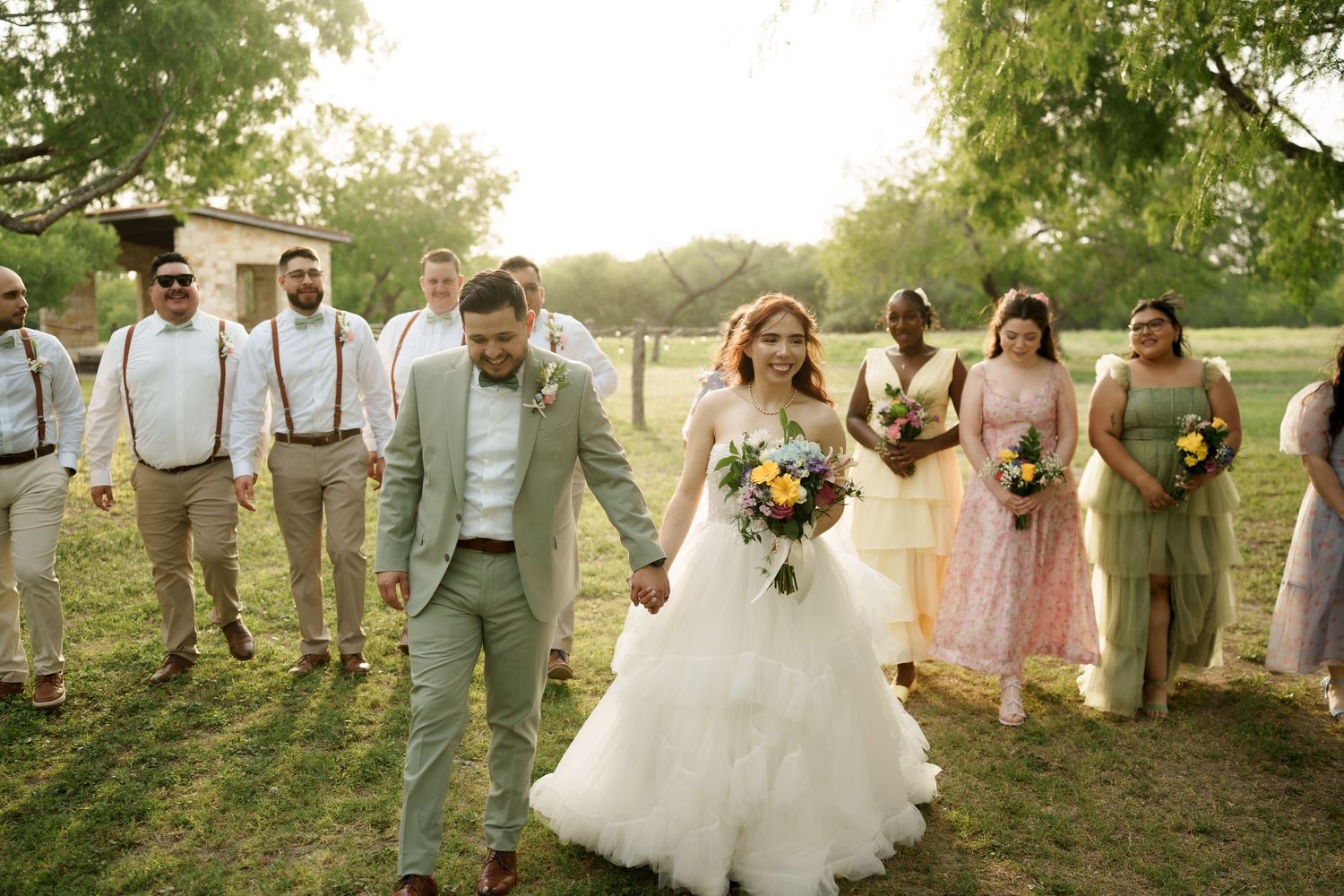 Wedding party in pastel attire walks through a green outdoor setting.