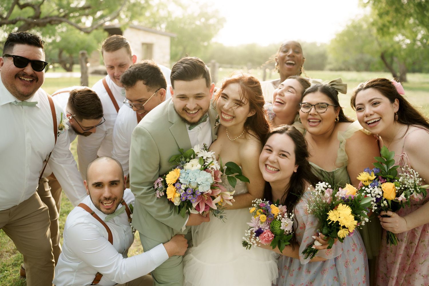 Groomsmen in light colored suits and suspenders pose together outdoors at a wedding celebration.