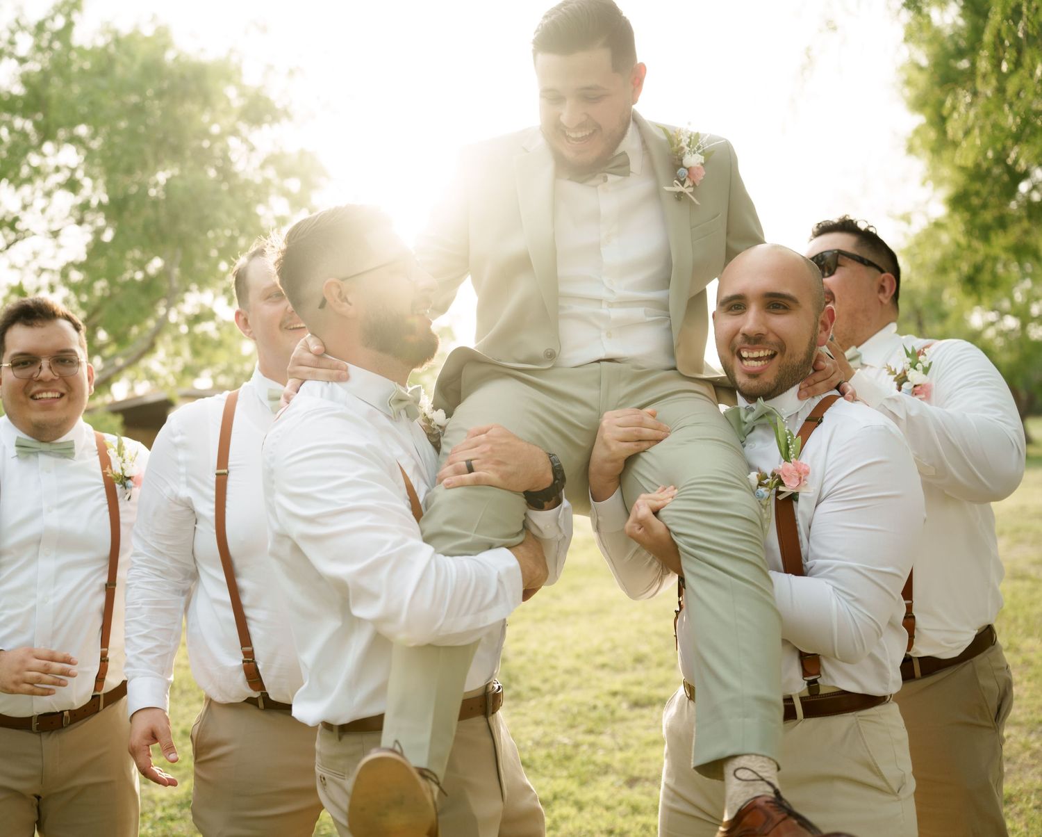 Wedding party members in suspenders and light colored attire laugh together in golden sunlight.