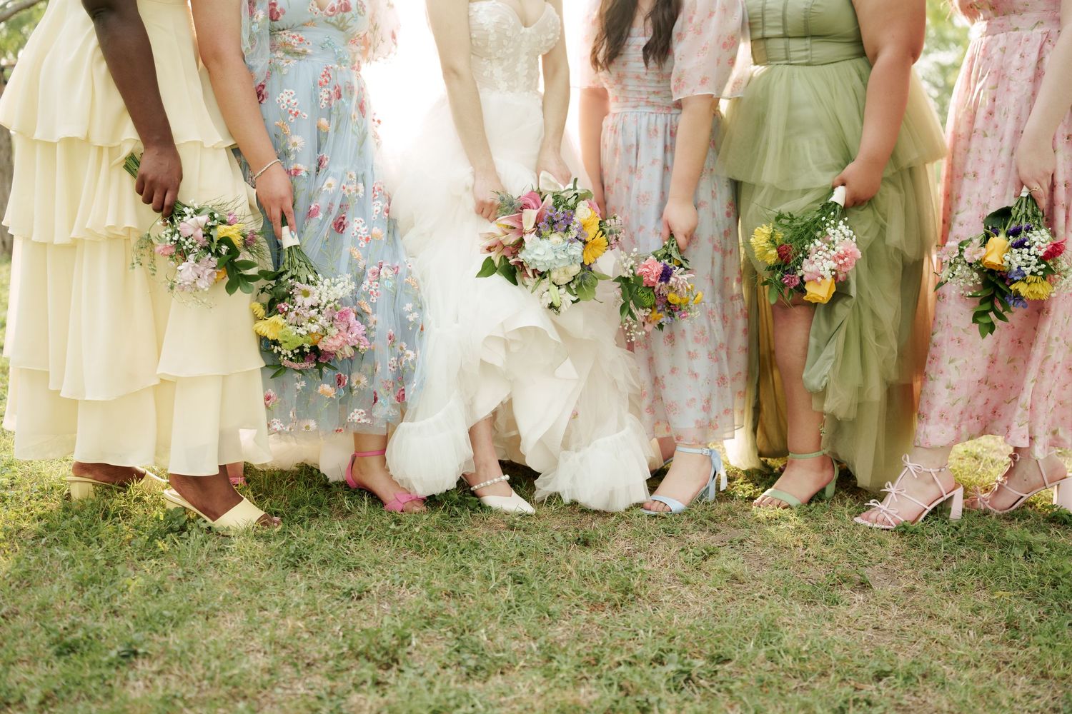 Row of bridesmaids' dresses in soft pastel colors shown from waist down at outdoor wedding.