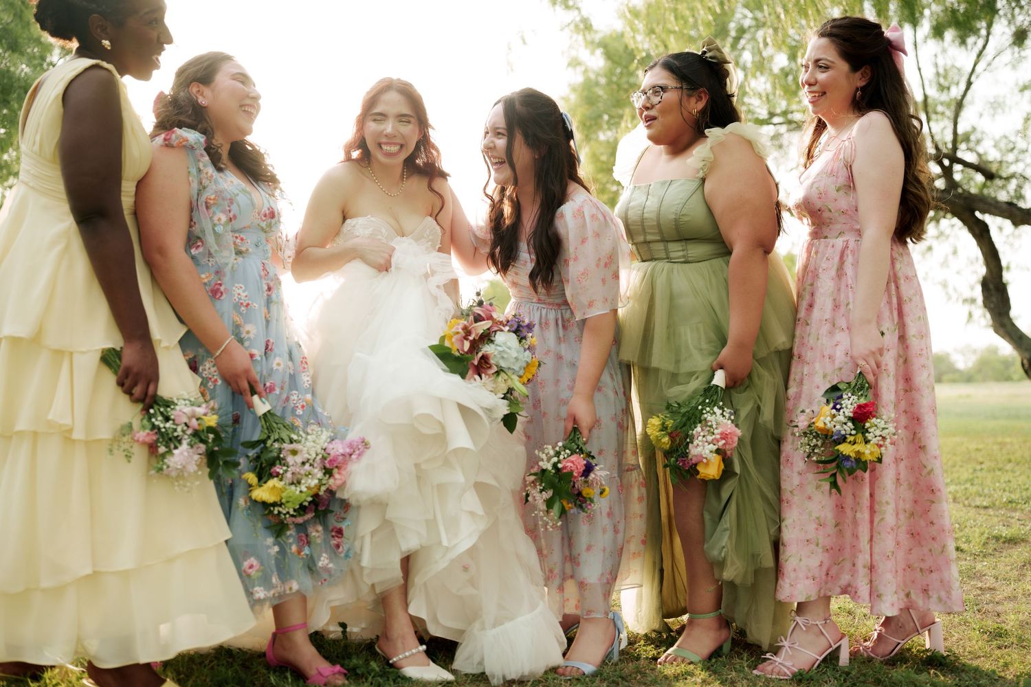 Wedding party wearing mix of pastel dresses poses together on grassy lawn.