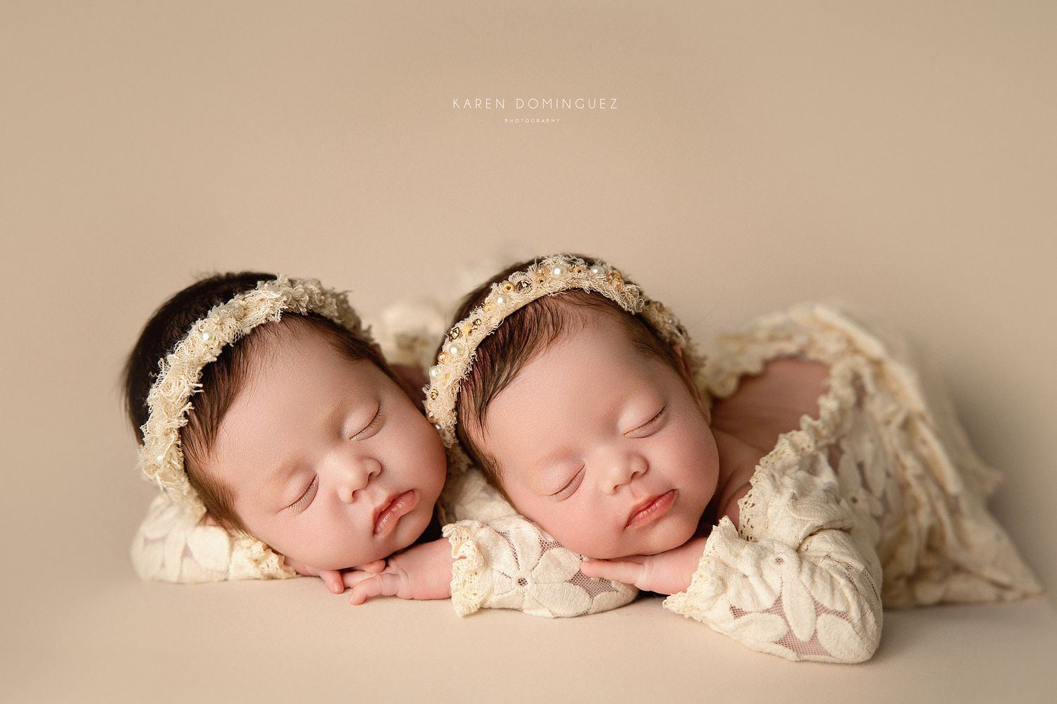 Two sleeping newborn babies wearing matching lace headbands lay side by side wrapped in cream-colored fabric.