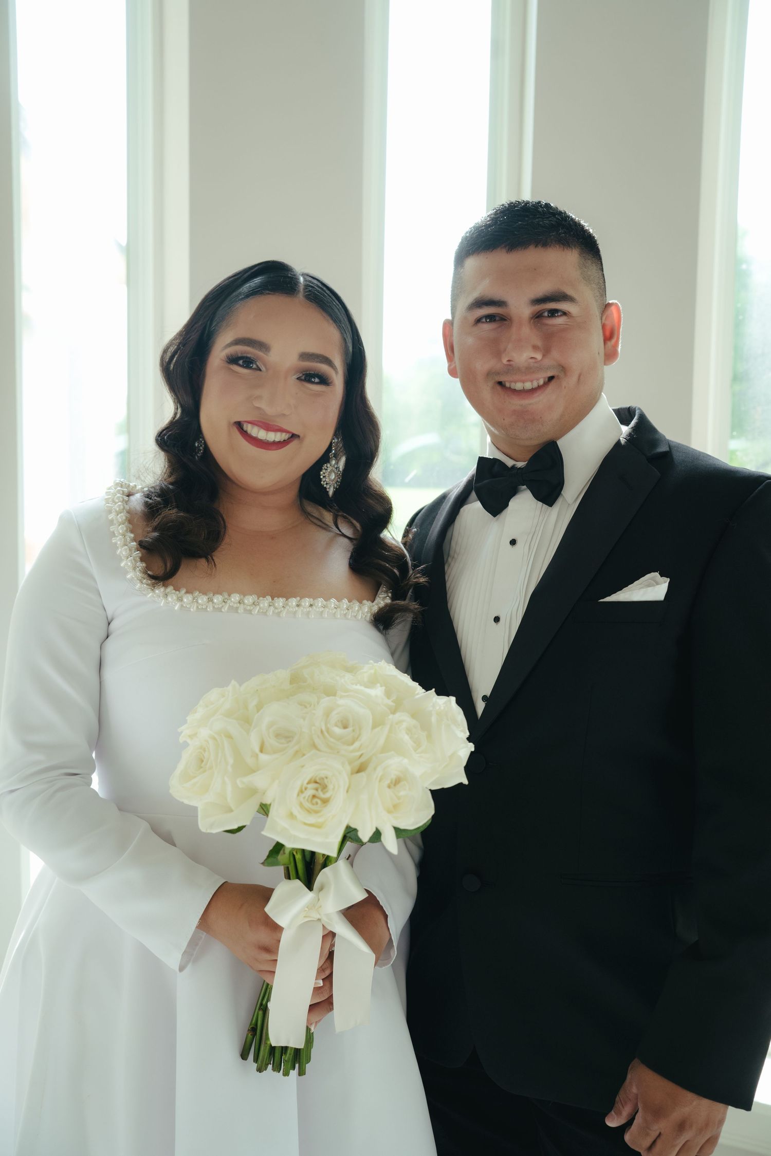 A wedding couple poses together in formal attire with a white rose bouquet.