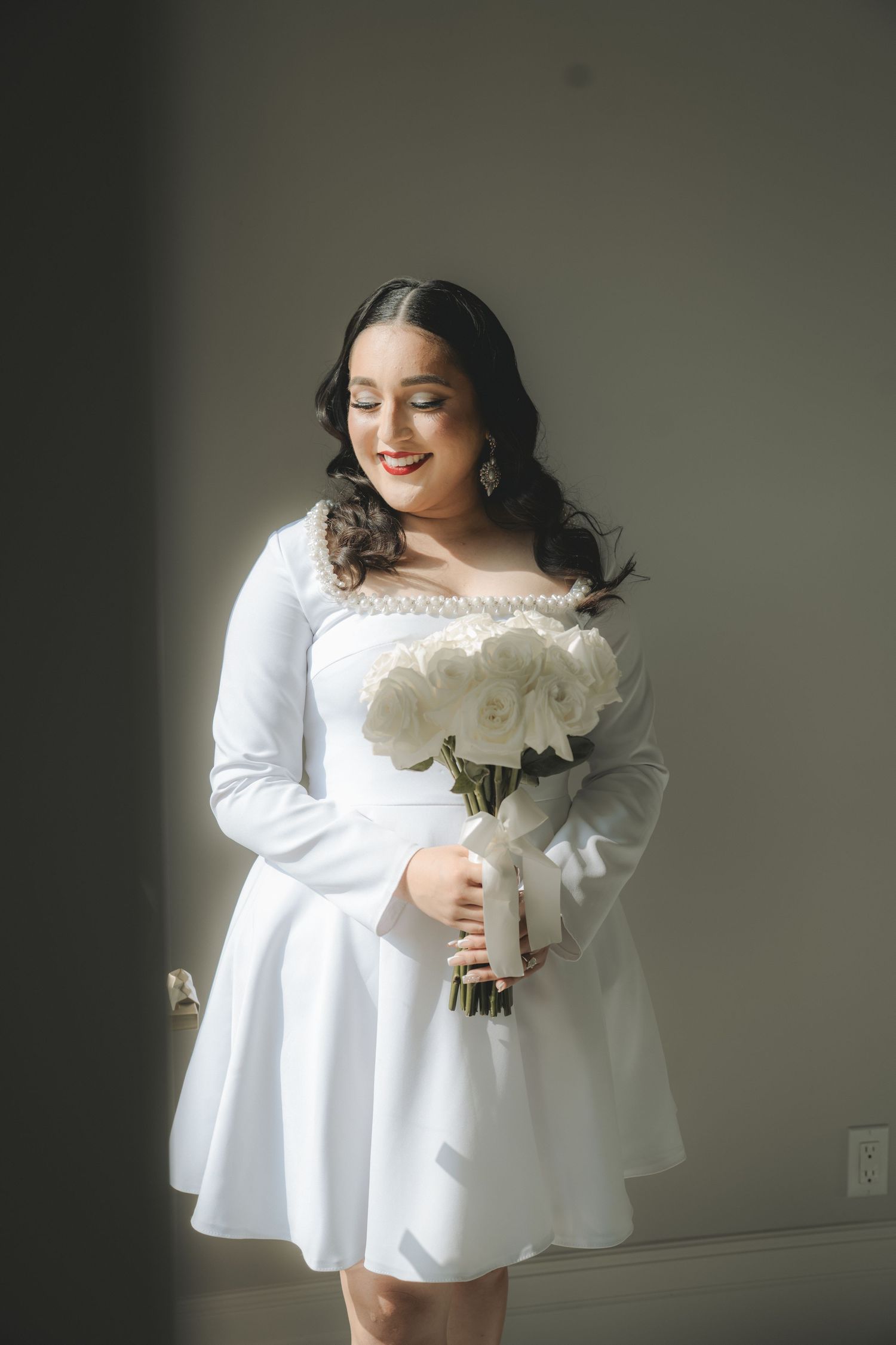 A bride in a white dress holds a classic white rose bouquet against a neutral background.