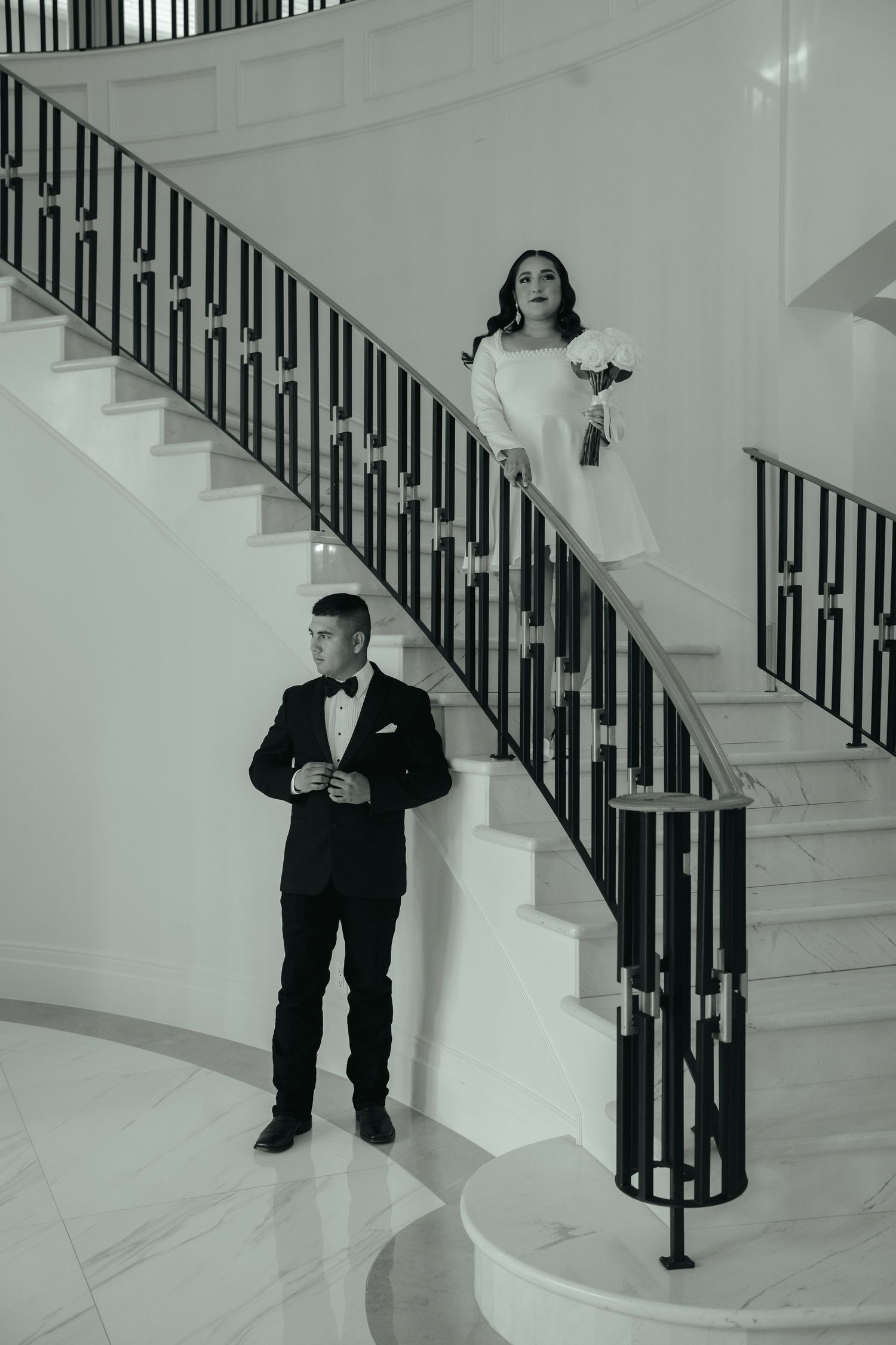 A couple in formal attire stands on an elegant white staircase with black railings.