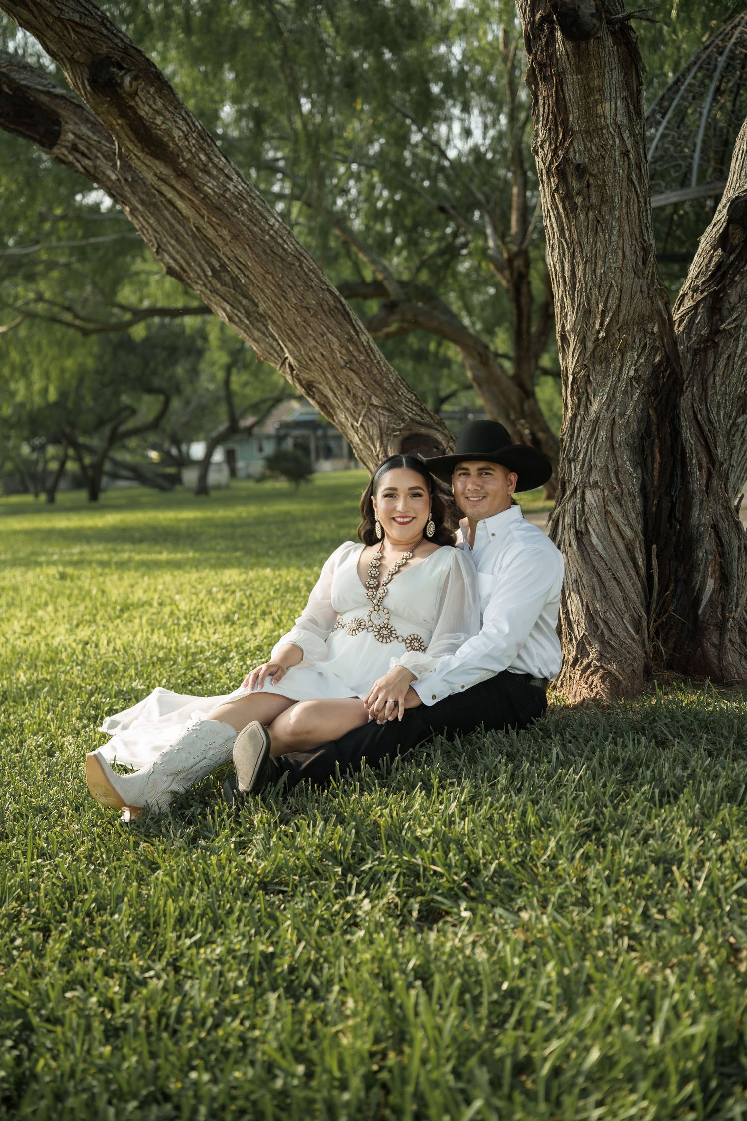 A romantic couple lounges under a large tree in a grassy field during a photo session.