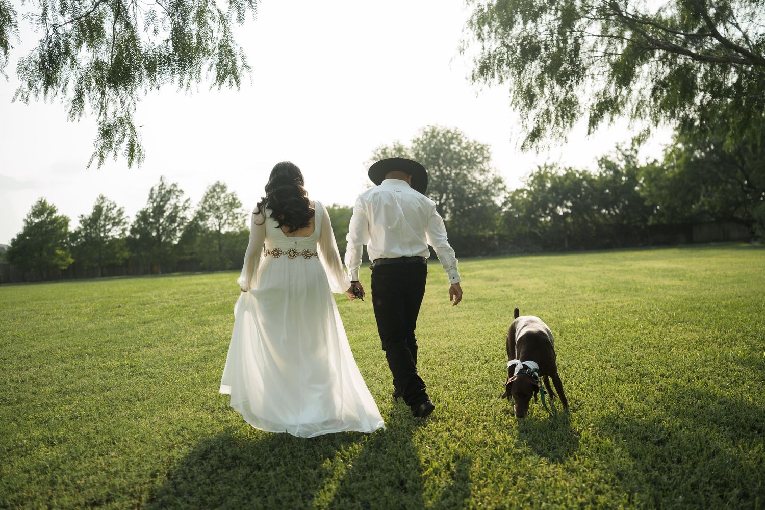 A couple walks hand in hand across a sunlit meadow in formal attire.