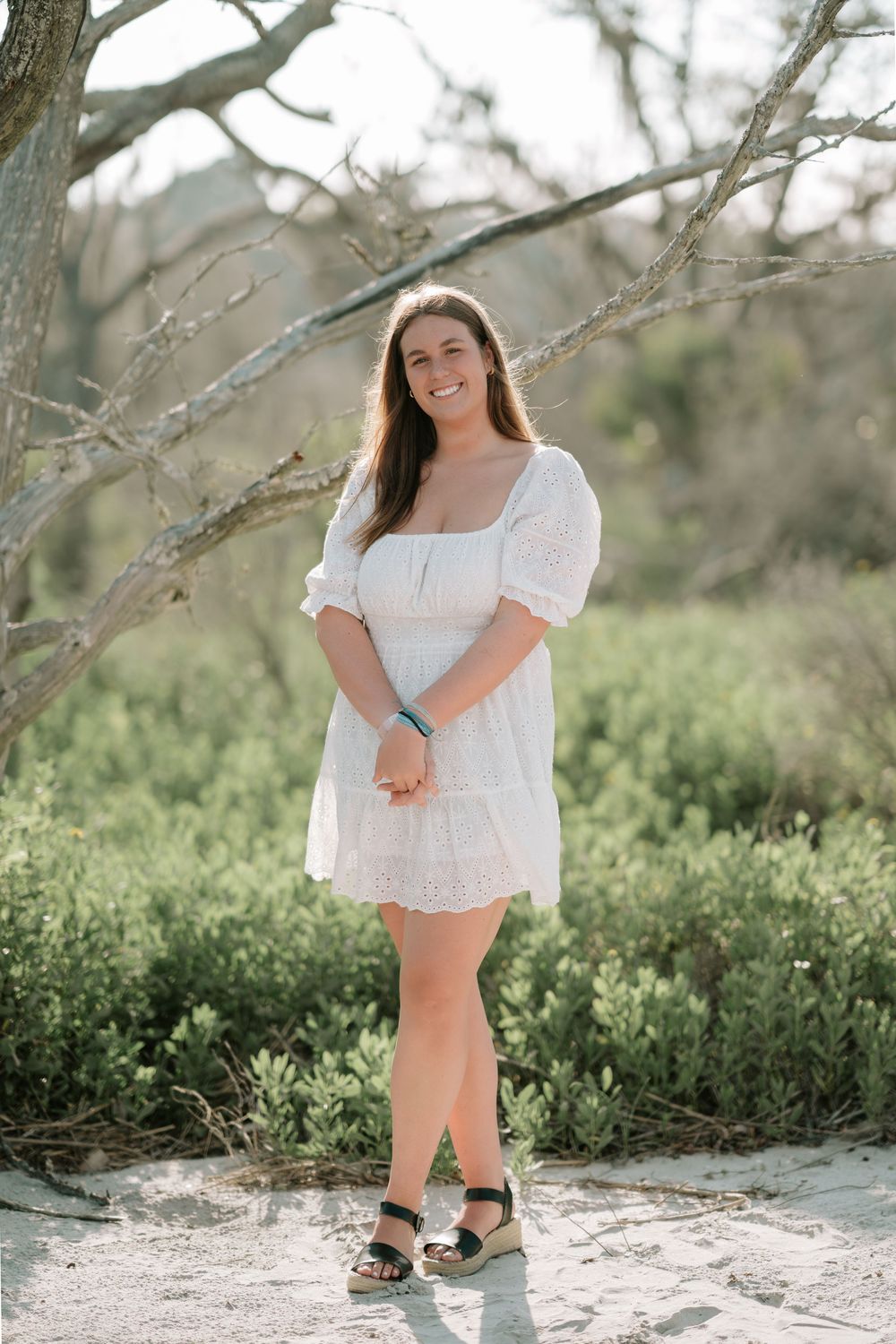High school senior girl posing barefoot among large driftwood trees on Jekyll Island beach at golden hour.